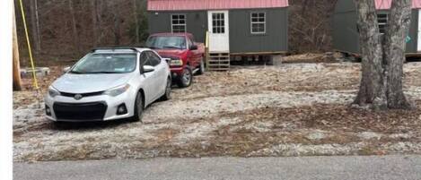 A small, two-story cabin with a red roof, a white car parked in front, and a red truck nearby.