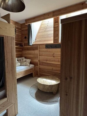 A wooden cabin interior with a loft bed, a round wooden stool, and a window with a view.