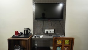 A room with a flat-screen TV mounted on a textured wall, a small wooden side table with a red mug, a kettle, and a remote control, and a colorful wooden chair. - Sarla Hotel Inn (Noida)