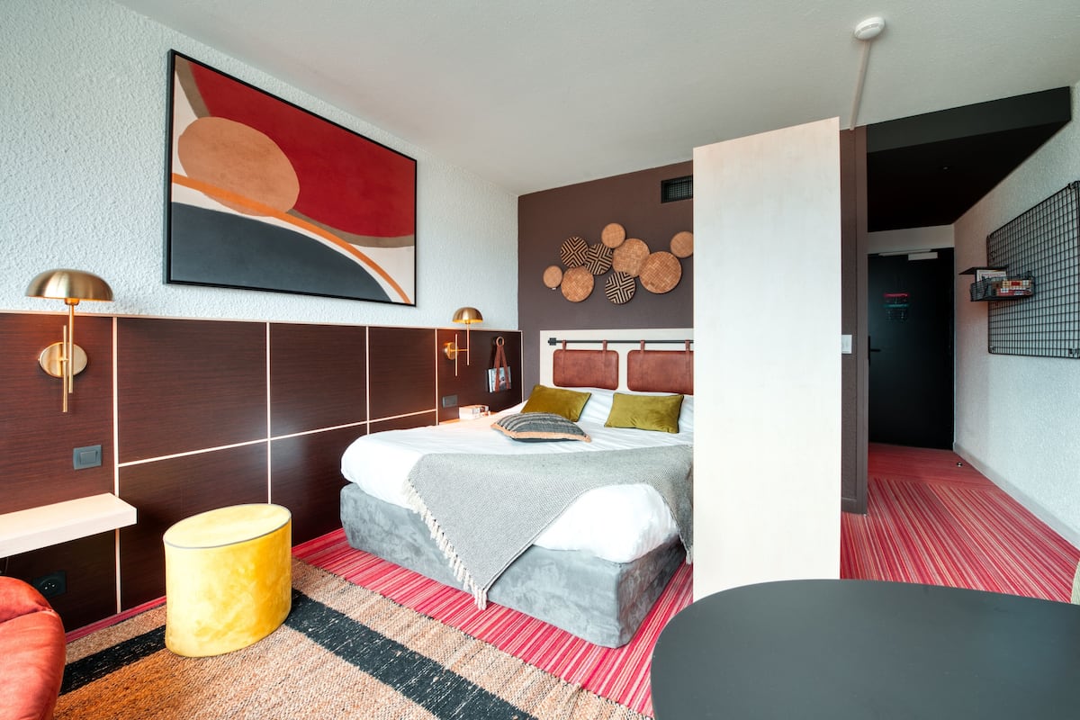 Habitación de hotel moderna con una cama grande, una mesita de noche, una lámpara, un otoman amarillo y una alfombra a rayas rojas.