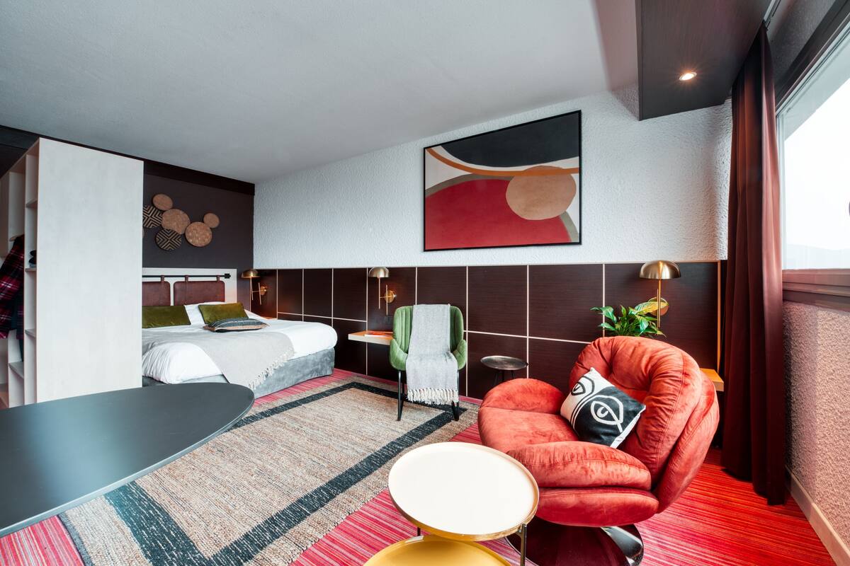 Una habitación de hotel moderna con una cama, un sillón rojo, una mesita y un cuadro abstracto grande en la pared.