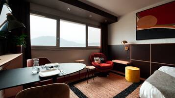 A modern hotel room with a large window, a round dining table, a red armchair, a yellow ottoman, and a bed with a striped blanket.