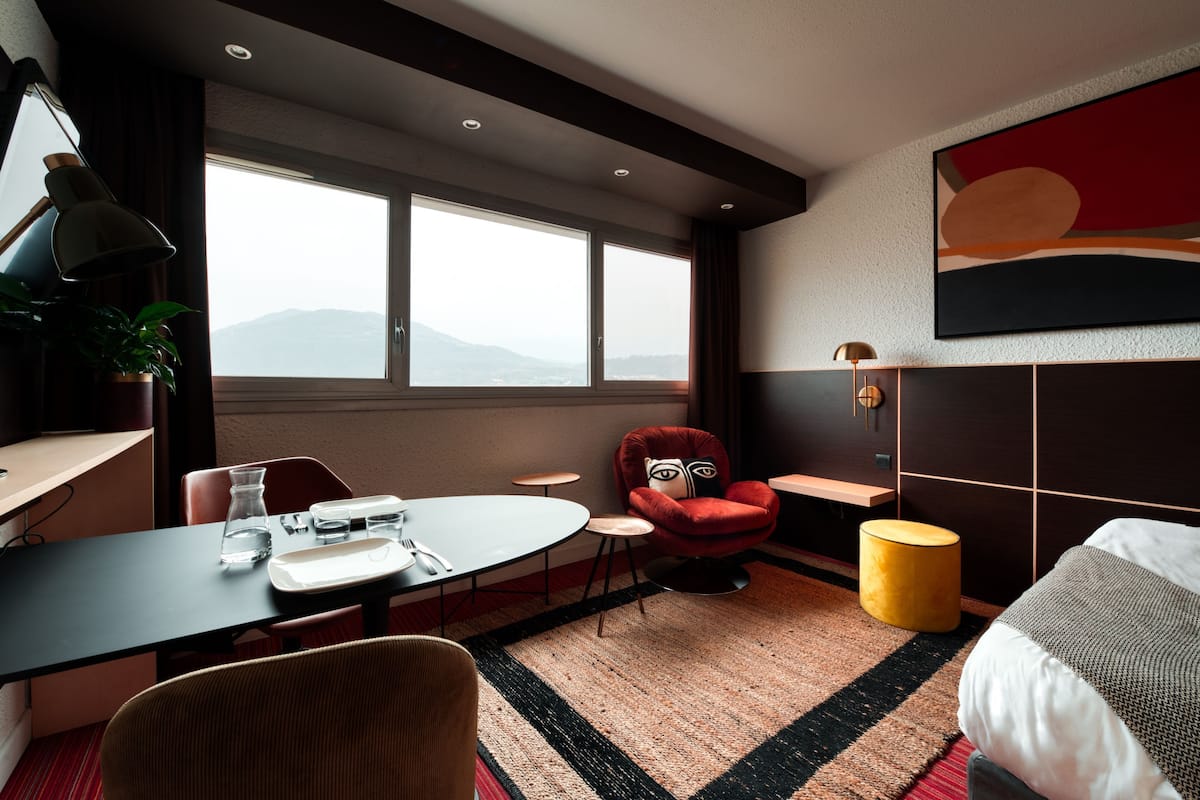 Habitación de hotel con cama, una mesa de comedor con sillas, un sillón rojo, una mesita redonda, una lámpara y un gran ventanal con vistas a las montañas.
