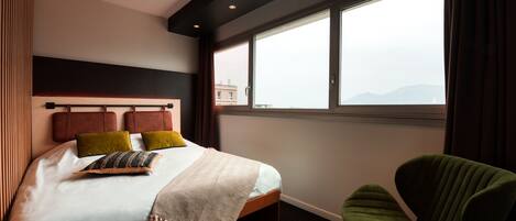 Quarto de hotel moderno com uma cama grande, uma cadeira verde e vista da cidade através de amplas janelas.