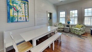Una habitación con una mesa blanca y un banco, dos sillones con estampado floral, una lámpara y un cuadro de palmeras.