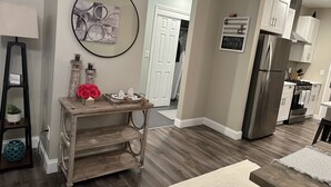 A modern kitchen with wooden flooring, white cabinets, a stainless steel refrigerator, and a wooden console table with decorative items. - Romantic Villa (Troy)