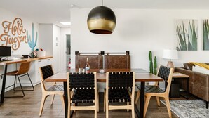 Een moderne eetruimte met een houten tafel, zwarte stoelen met kussens, een hanglamp en een wanddecoratie van een cactus.