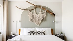 Een hotelkamer met een bed, een macramé wanddecoratie, een houten hoofdbord, een nachtkastje met een lamp en een bijzettafeltje met een klok en een plant.