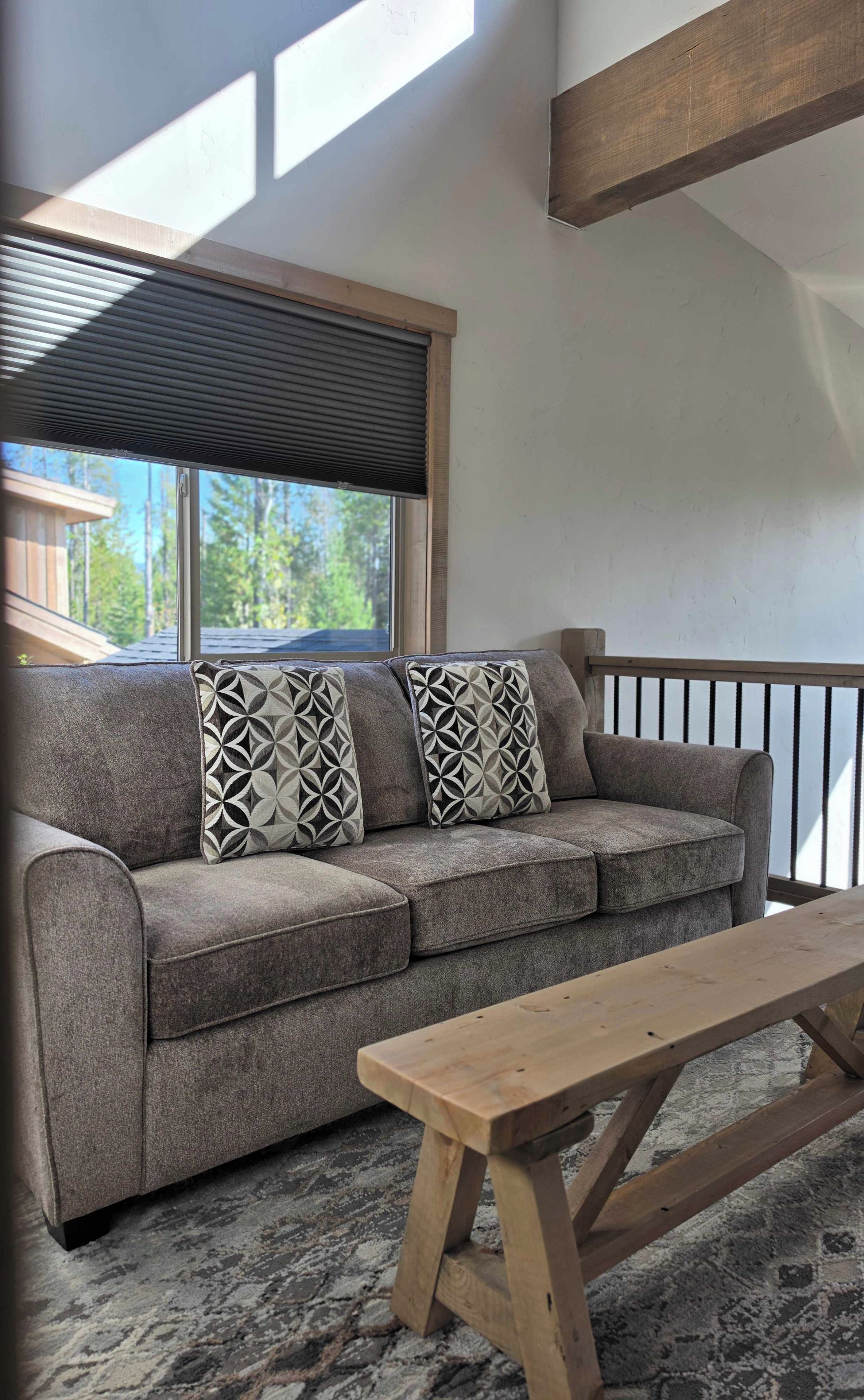 Un sofá gris con cojines estampados, un banco de madera y una ventana con persiana enrollable.