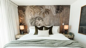 Un lit bien fait, recouvert d’une couverture verte, avec deux lampes de chevet et un mur décoré d’un motif de feuilles tropicales.