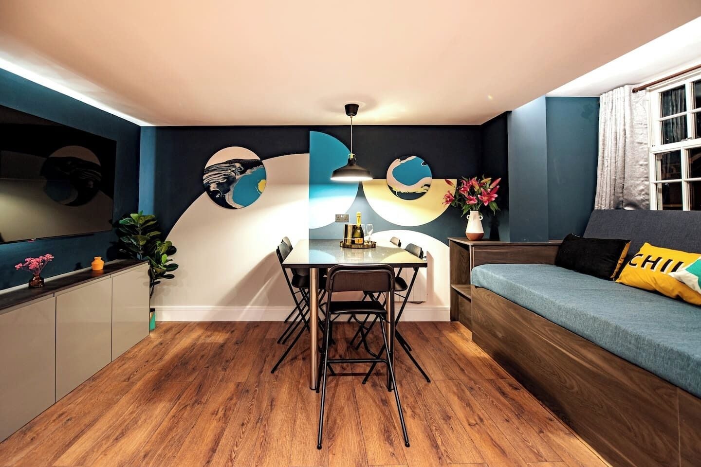A modern living space with a dining area, a sofa with a cushion, a table with chairs, and a wall with abstract art.