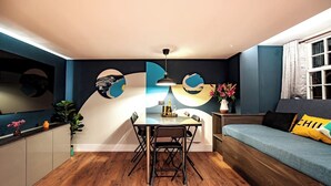 A modern living space with a dining area, a sofa with a cushion, a table with chairs, and a wall with abstract art. - The Nest: Luxury 16 person Apt w jacuzzi & parking (Blackpool)