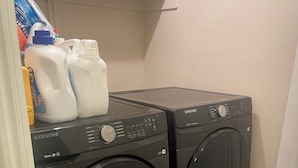 Un espace buanderie équipé de deux machines à laver Samsung, d’une étagère contenant des produits de nettoyage et d’un tapis à motifs sur le sol.