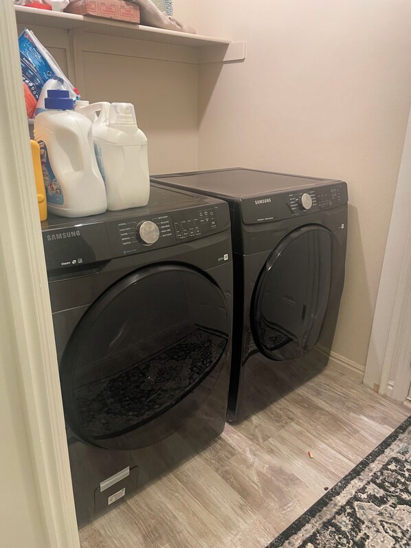 A laundry area with two Samsung washing machines, a shelf with cleaning supplies, and a patterned rug on the floor. - Quiet and comfortable home near SLC and ski resorts (North Salt Lake)