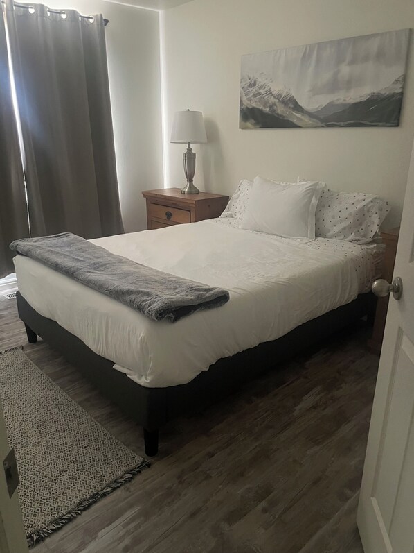 Una cama bien hecha con una manta gris, una mesita de noche de madera con una lámpara, una alfombra estampada y un cuadro de montañas en la pared.