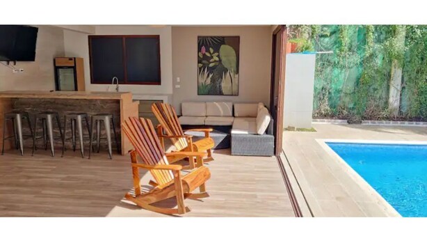 Una zona exterior moderna amb piscina, cadires de fusta, un sofà i una taula de dinar.