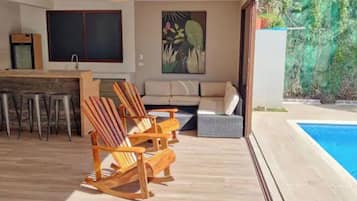 A modern outdoor living area with a pool, wooden chairs, a sofa, and a dining table.