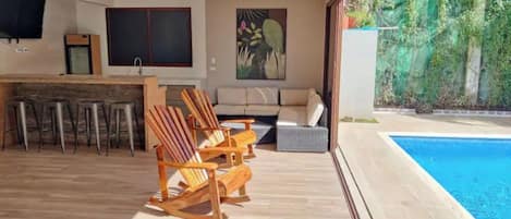 A modern outdoor living area with a pool, wooden chairs, a sofa, and a dining table.