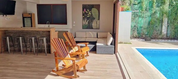 Una zona relax esterna moderna con piscina, sedie in legno, un divano e un tavolo da pranzo.