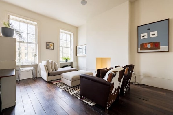 A living room with a sofa, ottoman, and a chair with a cowhide pattern. There are two windows, a plant, and a lamp on a side table. - Zone 1 RIBA Architect Designed home (London)