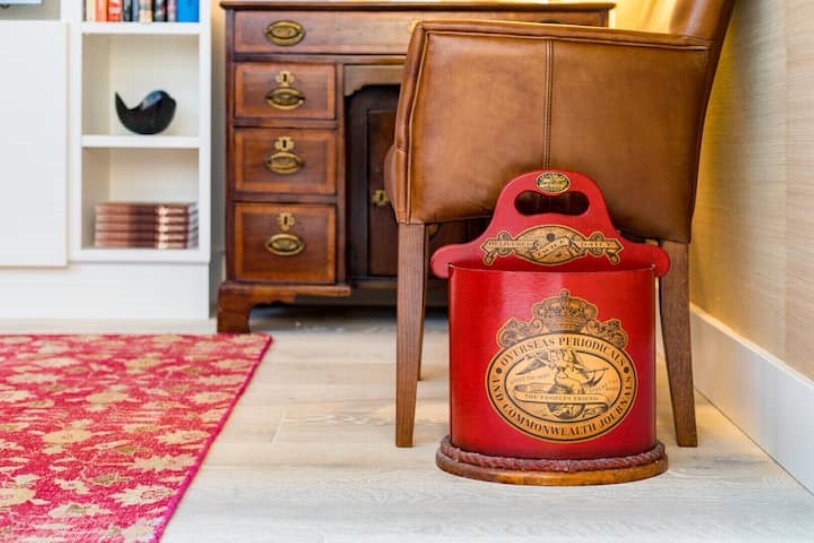 Un seau rouge orné d’un blason, posé sur un plancher en bois, à côté d’un fauteuil en cuir marron et d’un meuble en bois dont les poignées sont en laiton.