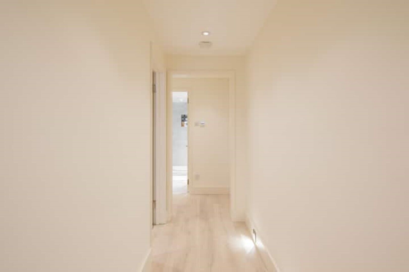 A narrow hallway with light-colored walls and wooden flooring, leading to a door at the end.