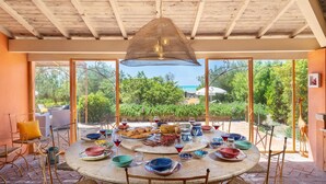 A round table set for dining with various dishes and drinks, surrounded by wicker chairs, under a wooden ceiling with a hanging light fixture. - Villa Corallina 12, Emma Villas (Marina di Bibbona)