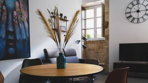 A modern living room with a round wooden table, black and brown chairs, a blue vase with dried plants, a large abstract painting, a stone wall, and a black clock. - Elegante Suite a piano terra di un palazzo storico del 1700 - Centro di Ortigia. (Siracusa)