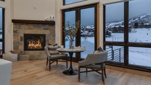 Una habitación acogedora con chimenea de piedra, una mesa redonda con sillas y vistas a un paisaje nevado a través de amplios ventanales.