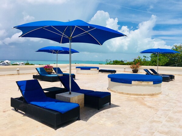 Un área de descanso en la playa con sombrillas azules, sillones reclinables y una piscina.