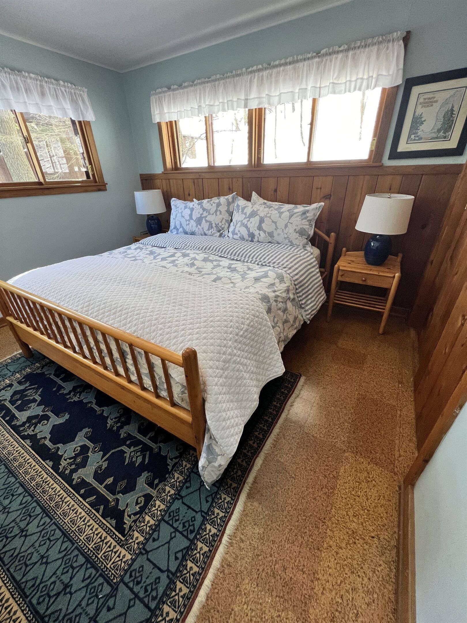 Un dormitorio con una cama de madera, dos mesitas de noche con lámparas, una alfombra estampada y una ventana con un visillo blanco.