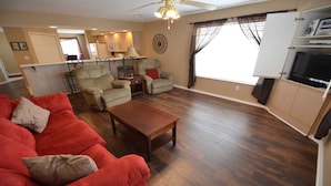 Een woonkamer met een rode bank, een houten salontafel en twee beige fauteuils. In de achtergrond is een keuken te zien met een koelkast, magnetron en kastjes.