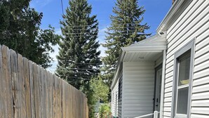 Ein schmaler Pfad führt zu einem Haus, an dessen Seite ein Holzzaun und auf der anderen Seite ein Garten mit einem großen Pflanzgefäß zu sehen sind.