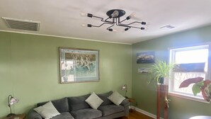 Ein Wohnzimmer mit einem grauen Ecksofa, einer Deckenleuchte, einem gerahmten Bild und einem Fenster mit Pflanzen.