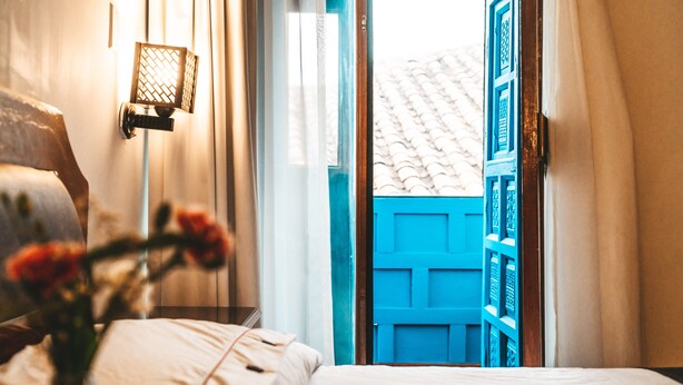 Habitación de hotel con cama, mesita de noche con lámpara y vista a una puerta azul y paredes blancas.