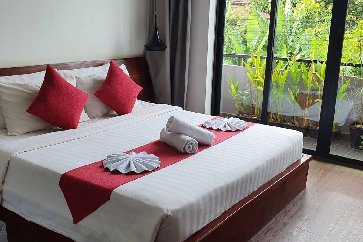 Un lit bien fait, avec des draps blancs et des oreillers rouges, un plaid rouge et deux serviettes pliées sur un tapis rouge assorti.