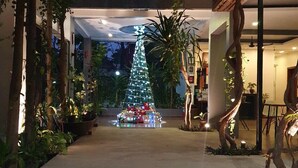 Een goed verlichte binnenruimte met een grote kerstboom, omringd door kamerplanten en een trap die naar een hoger gelegen verdieping leidt.