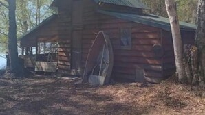 Una cabaña rústica de troncos con una noria grande, rodeada de árboles.