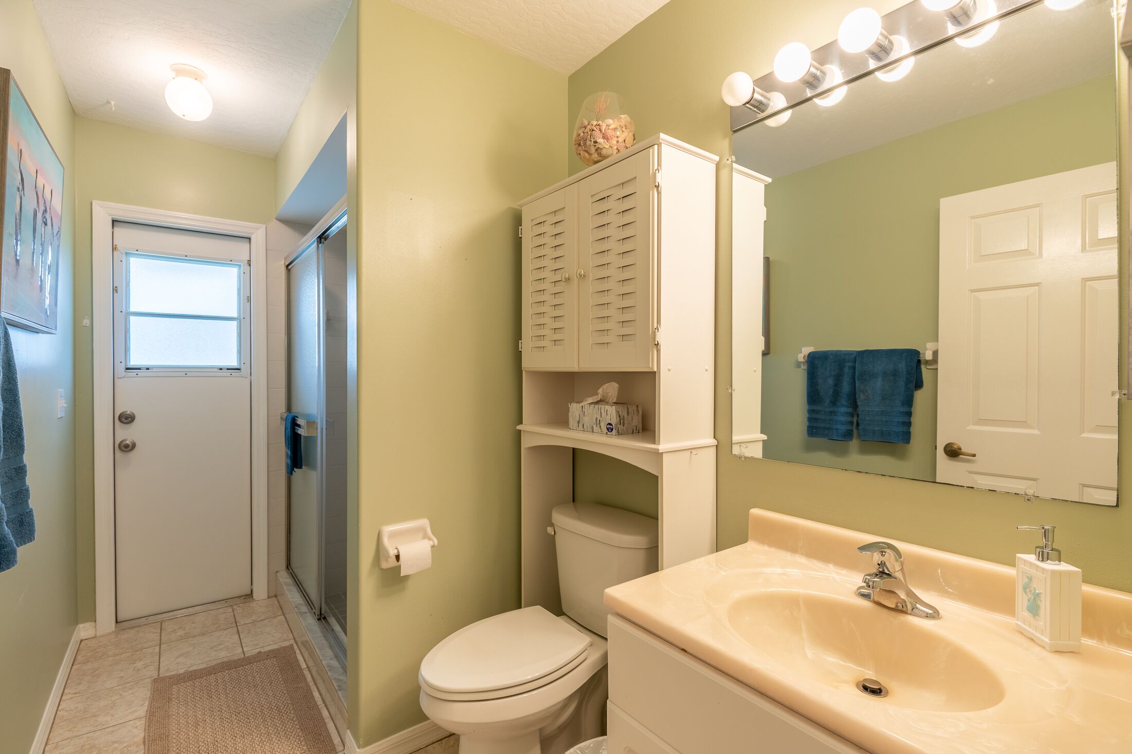 A bathroom with a toilet, sink, and mirror. There is a window, a towel rack, and a cabinet.