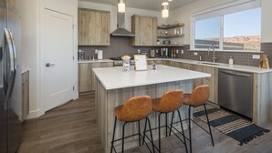 A modern kitchen with wooden cabinets, a central island with bar stools, a built-in oven, and a large window offering a desert view. - Magnificent 4 bedroom New home with views minutes from Arches (Moab)