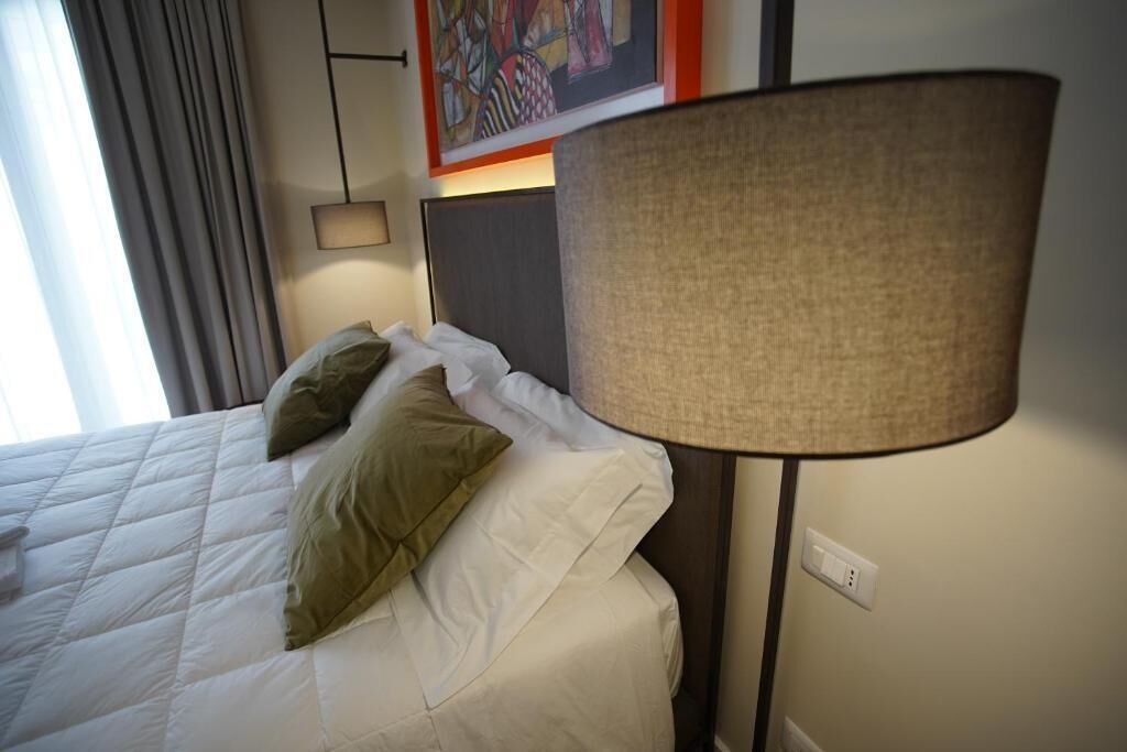 A neatly made bed with white and green pillows, a dark headboard, and a large floor lamp.