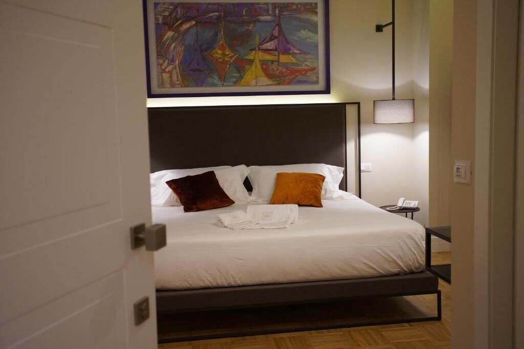 Cama bem arrumada com lençóis brancos e duas almofadas, mesa de cabeceira com abajur, e um quadro sobre a cama.