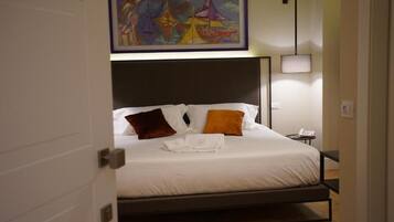 Cama bem arrumada com lençóis brancos e duas almofadas, mesa de cabeceira com abajur, e um quadro sobre a cama.