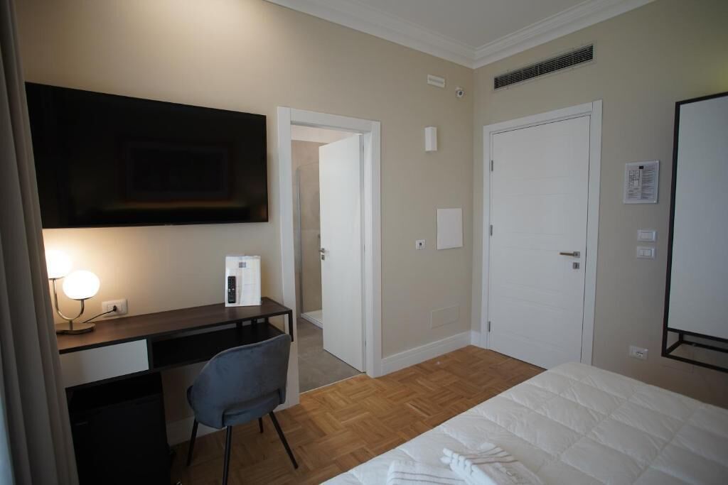 A hotel room with a bed, a desk with a chair, a TV mounted on the wall, and a door leading to another room.