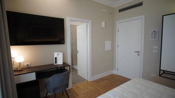 A hotel room with a bed, a desk with a chair, a TV mounted on the wall, and a door leading to another room.