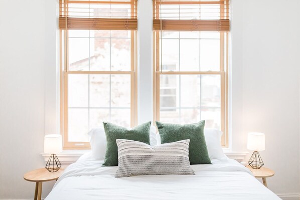 A neatly made bed with green and striped pillows, two bedside tables with lamps, and large windows with blinds.