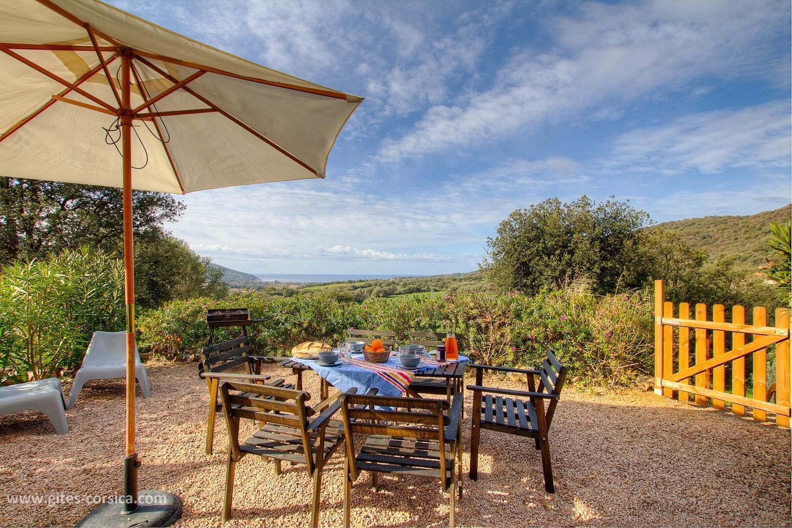 An outdoor dining area with a table set for two, surrounded by wooden chairs and a large umbrella.
