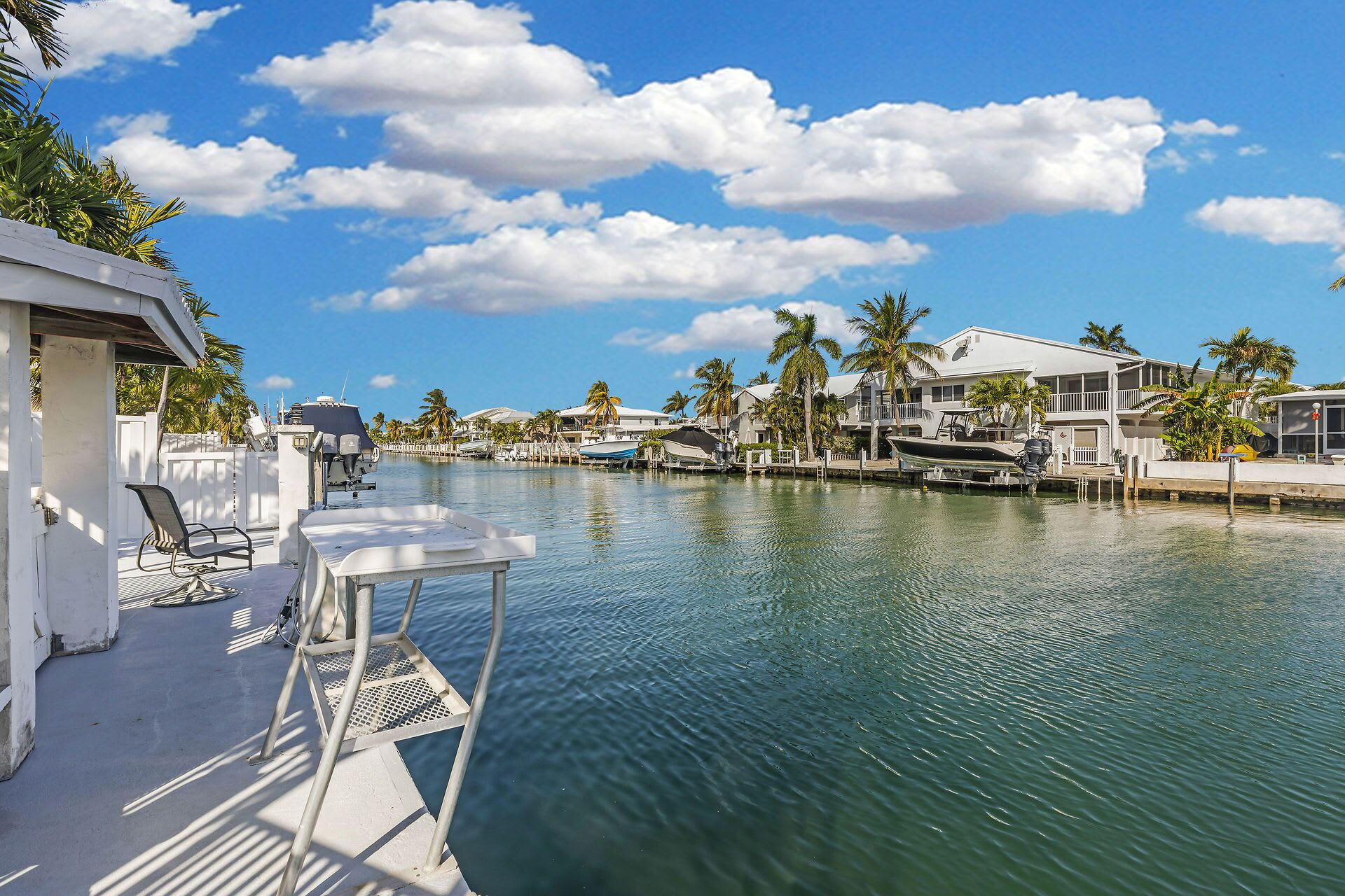 A marina with boats docked, a pool area with lounge chairs, and houses with palm trees in the background.