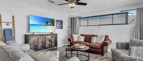 A living room with a ceiling fan, a television displaying a beach scene, a brown leather sofa, and patterned armchairs.