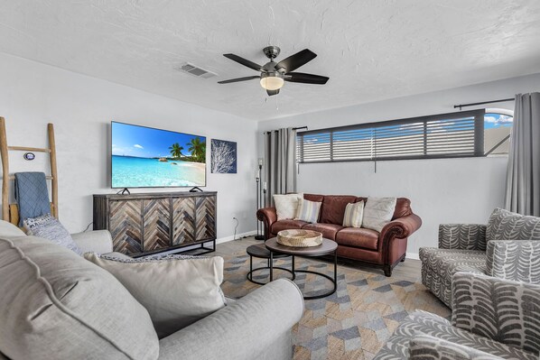 A living room with a ceiling fan, a television displaying a beach scene, a brown leather sofa, and patterned armchairs.